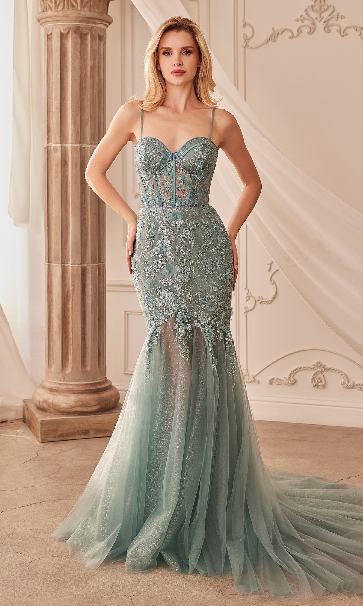 A woman stands indoors wearing the Andrea and Leo A1232 long lace mermaid prom dress in light blue, featuring sheer panels and a tulle skirt—an elegant choice for any special evening.